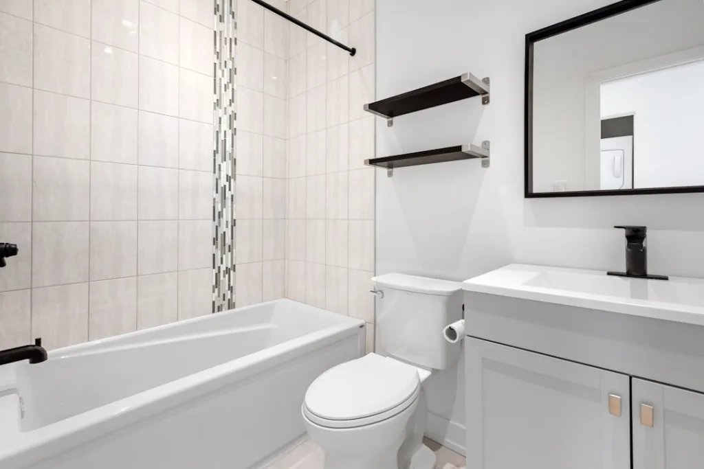 Small bathroom with a white vanity, modern fixtures, tiled walls, and floating shelves, illustrating cost-effective renovation elements.