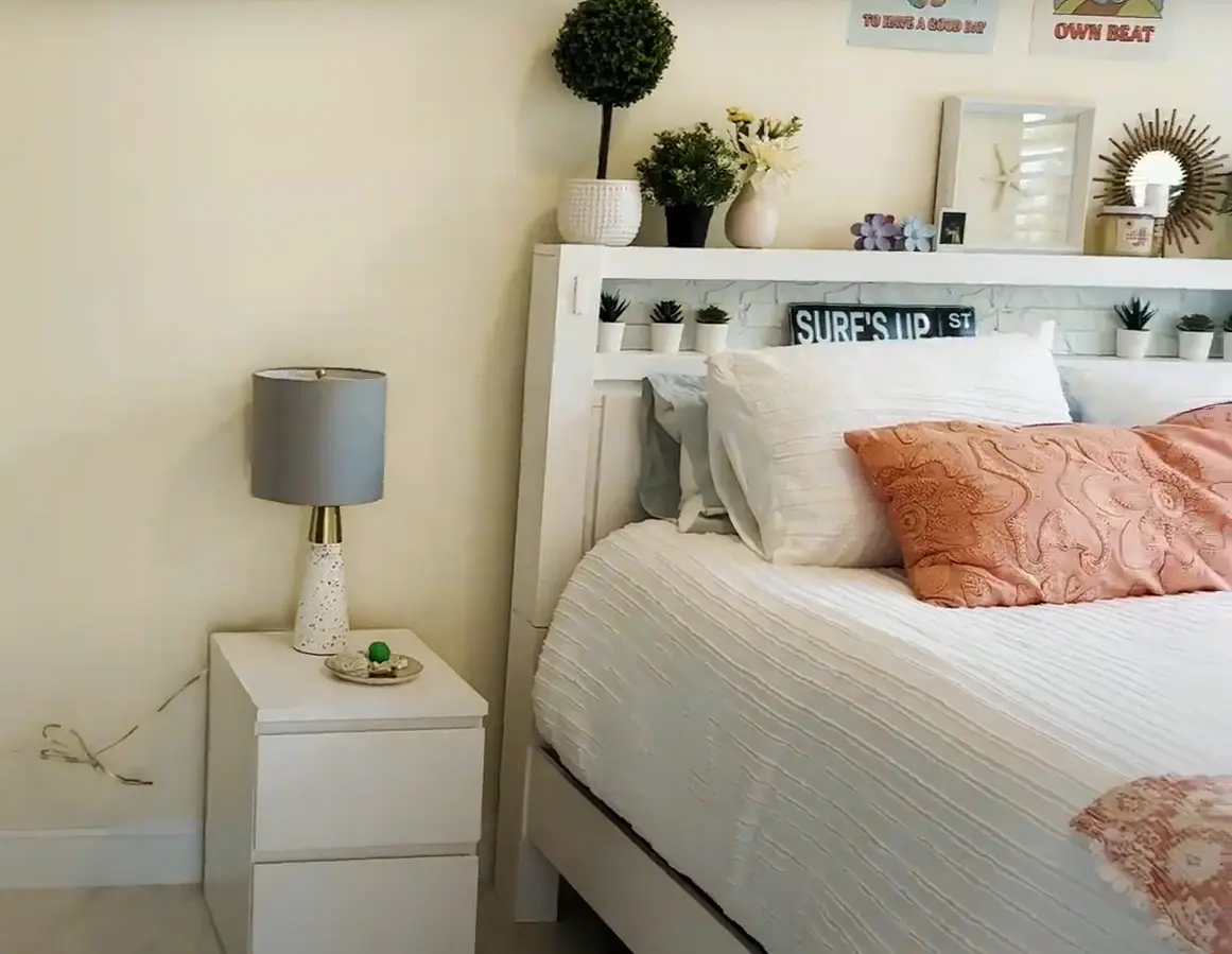 A remodeled bedroom featuring a neatly made bed with decorative pillows, a modern nightstand with a lamp, and a stylish shelf with plants and decor.