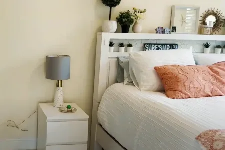 A remodeled bedroom featuring a neatly made bed with decorative pillows, a modern nightstand with a lamp, and a stylish shelf with plants and decor.